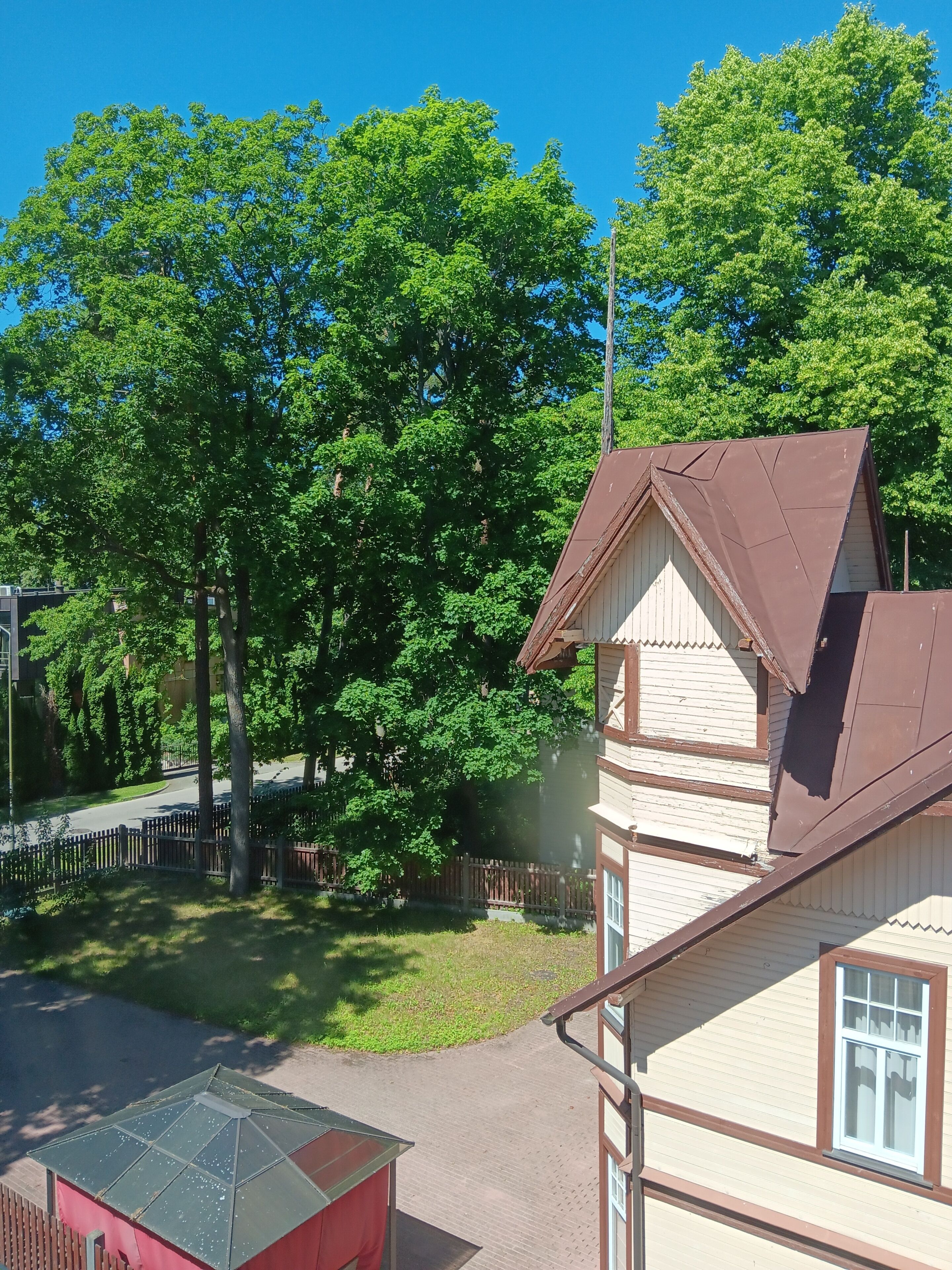 A residential area with a house featuring a brown roof and white siding, surrounded by trees and a fenced yard.