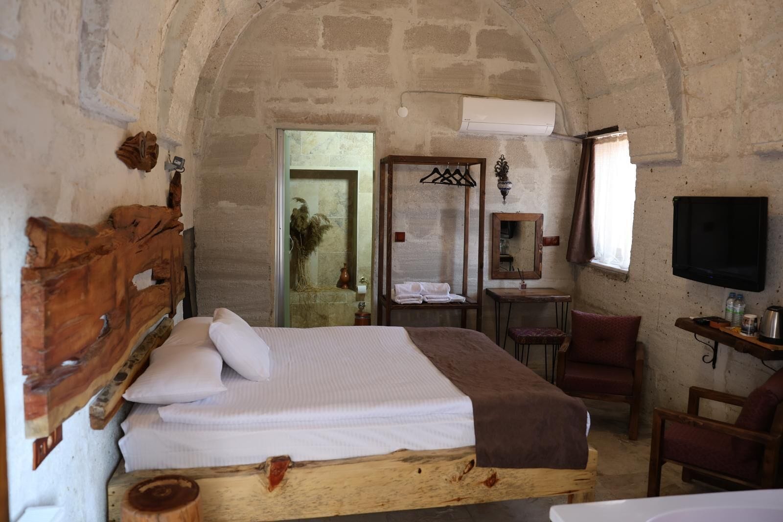 A rustic room with a wooden bed, a small desk, a mirror, a TV, and a shower area.
