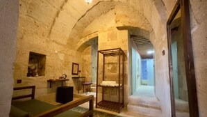 A stone-walled room with a bed, a wooden bench, a small table, and a hanging rack. - Almula Cave Hotel (Avanos)