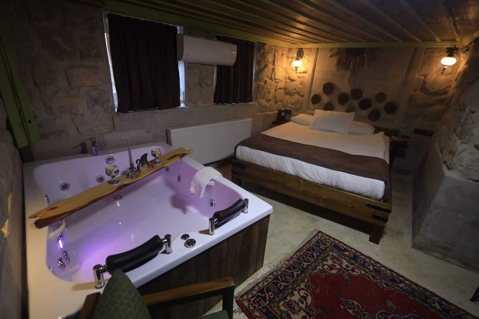 A rustic room with a stone wall, a wooden bed, a large bathtub with a wooden shelf, and a red patterned rug.
