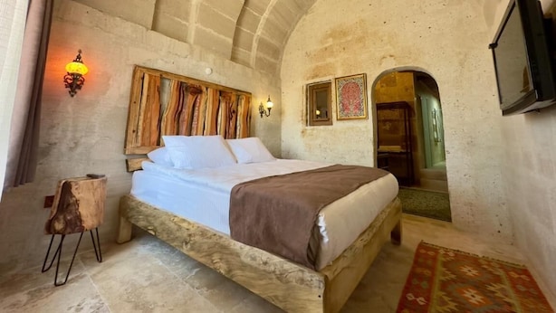 A bedroom with a stone ceiling, a bed with white linens, a wooden headboard, and a small wooden stool. - Almula Cave Hotel (Avanos)