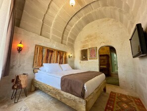 A bedroom with a stone ceiling, a bed with white linens, a wooden headboard, and a small wooden stool. - Almula Cave Hotel (Avanos)