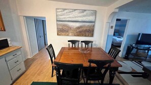 Un comedor con mesa y sillas de madera, un cuadro en la pared y vista a otra habitación.
