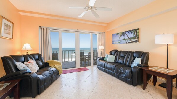 A living room with two black leather sofas, a ceiling fan, and a large window with a view of the ocean.