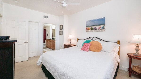 A bedroom with a bed, bedside tables, a ceiling fan, a bathroom visible through an open door, and a beach-themed picture on the wall.