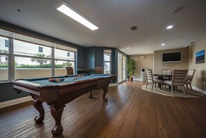 A pool table in a room with large windows, a dining area, and a flat-screen TV on the wall.