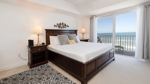 A bedroom with a large bed, two bedside tables, a nightstand, a patterned rug, and a view of the ocean through a balcony.