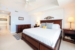 A bedroom with a large bed, a ceiling fan, a nightstand with a lamp, a dresser, and a rug.