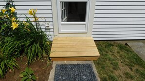 Eine Holzterrasse führt zu einer weißen Tür mit Fensterglas, umgeben von grünen Pflanzen und gelben Blumen.