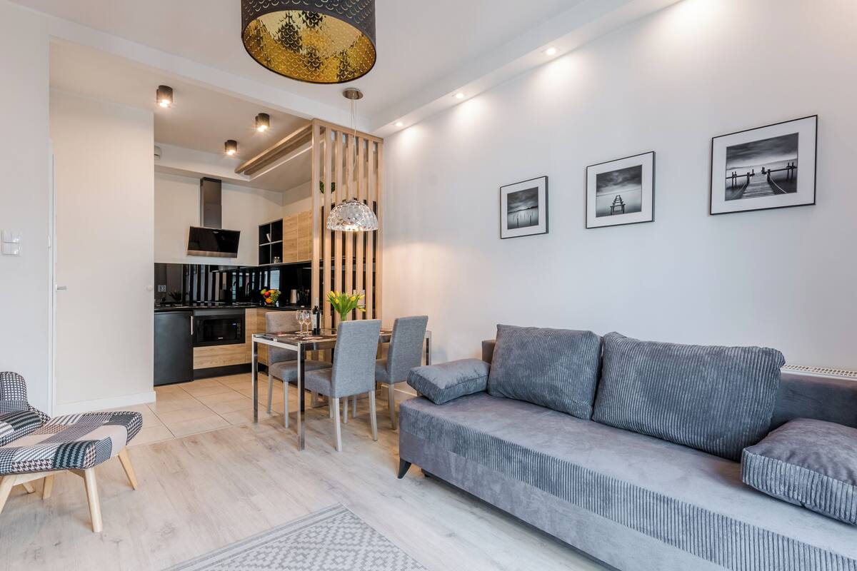 A modern living room with a grey sofa, dining table, and chairs. There is a kitchen area in the background with a TV and wooden cabinets.