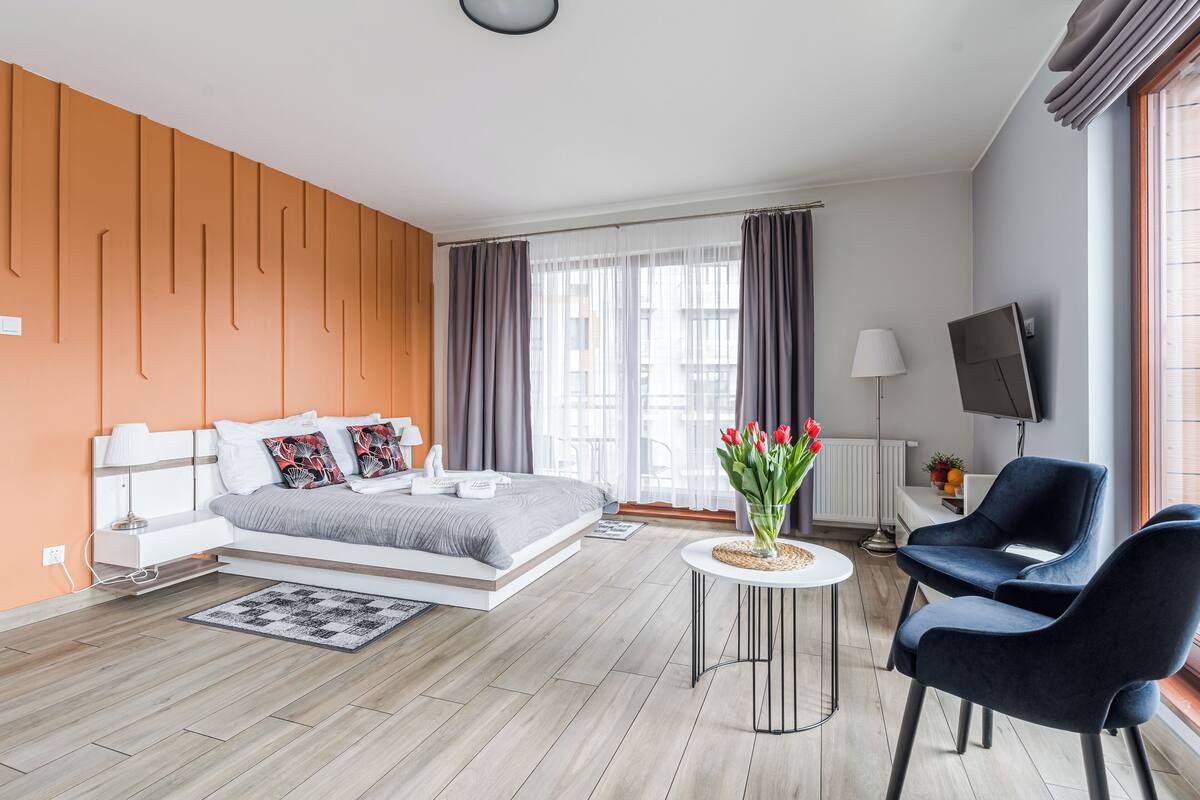 A modern hotel room with a bed, a round table, two chairs, a TV, and a window with curtains.