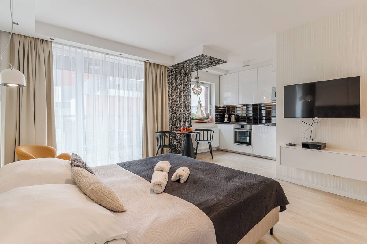 A modern hotel room with a large bed, a TV, a dining area, and a kitchenette.