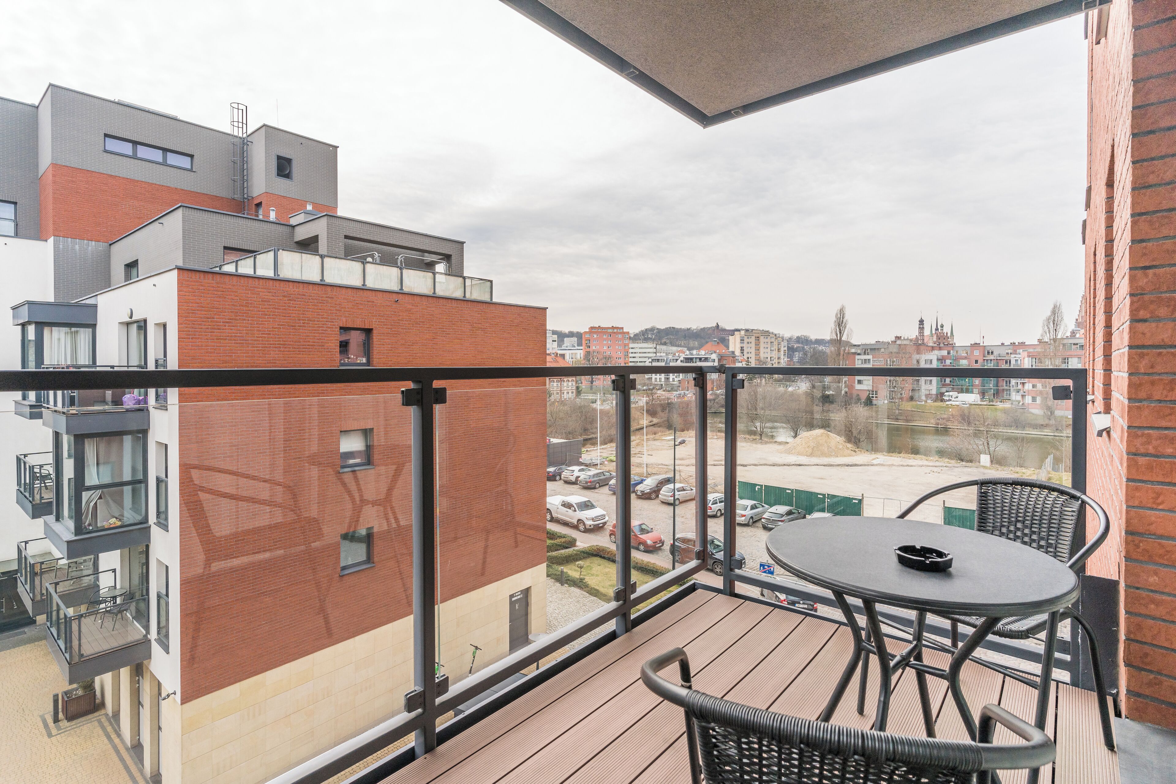 A balcony with a table and chairs, overlooking a parking area and buildings.