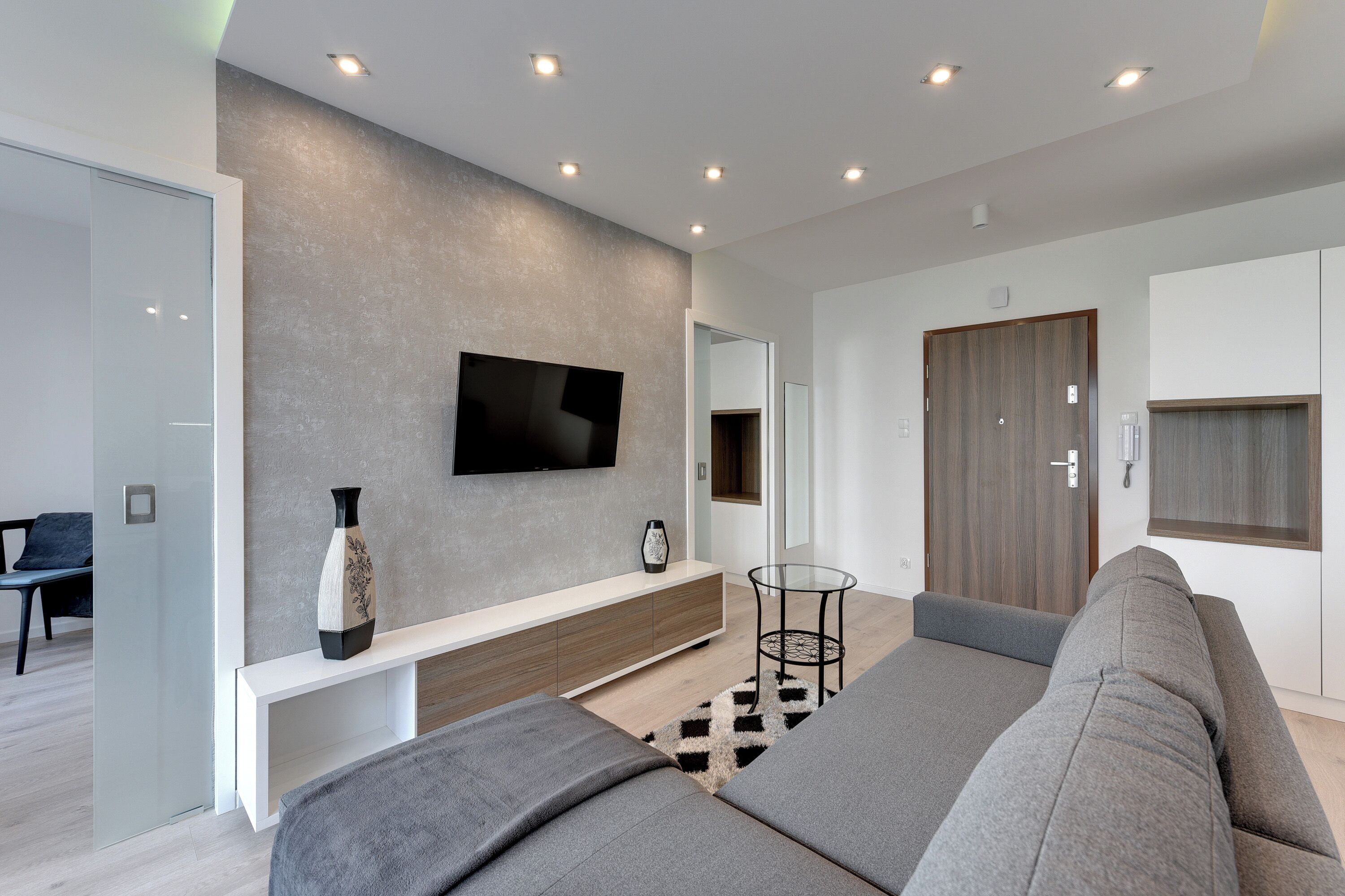 A modern living room with a grey sofa, a white console table, a flat-screen TV mounted on a grey textured wall, and a wooden door.