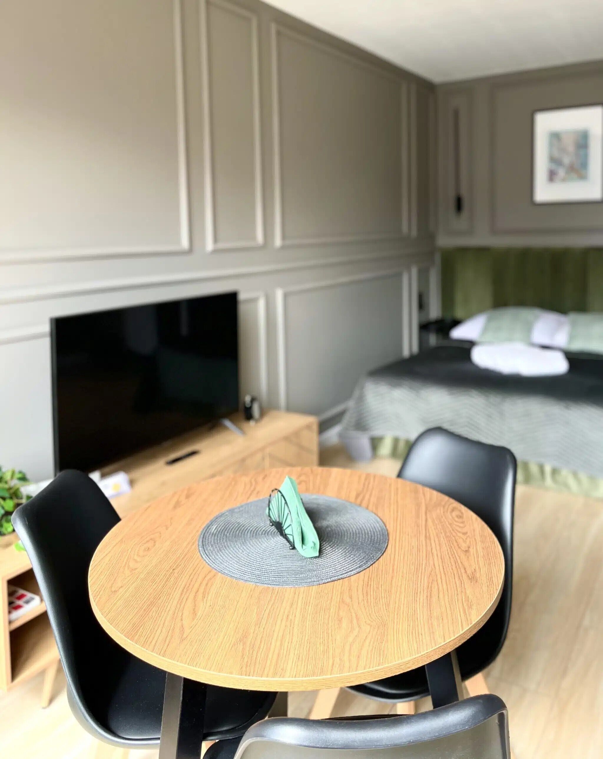 Een moderne kamer met een ronde houten tafel, zwarte stoelen, een televisie, een bed en een groen kussen.