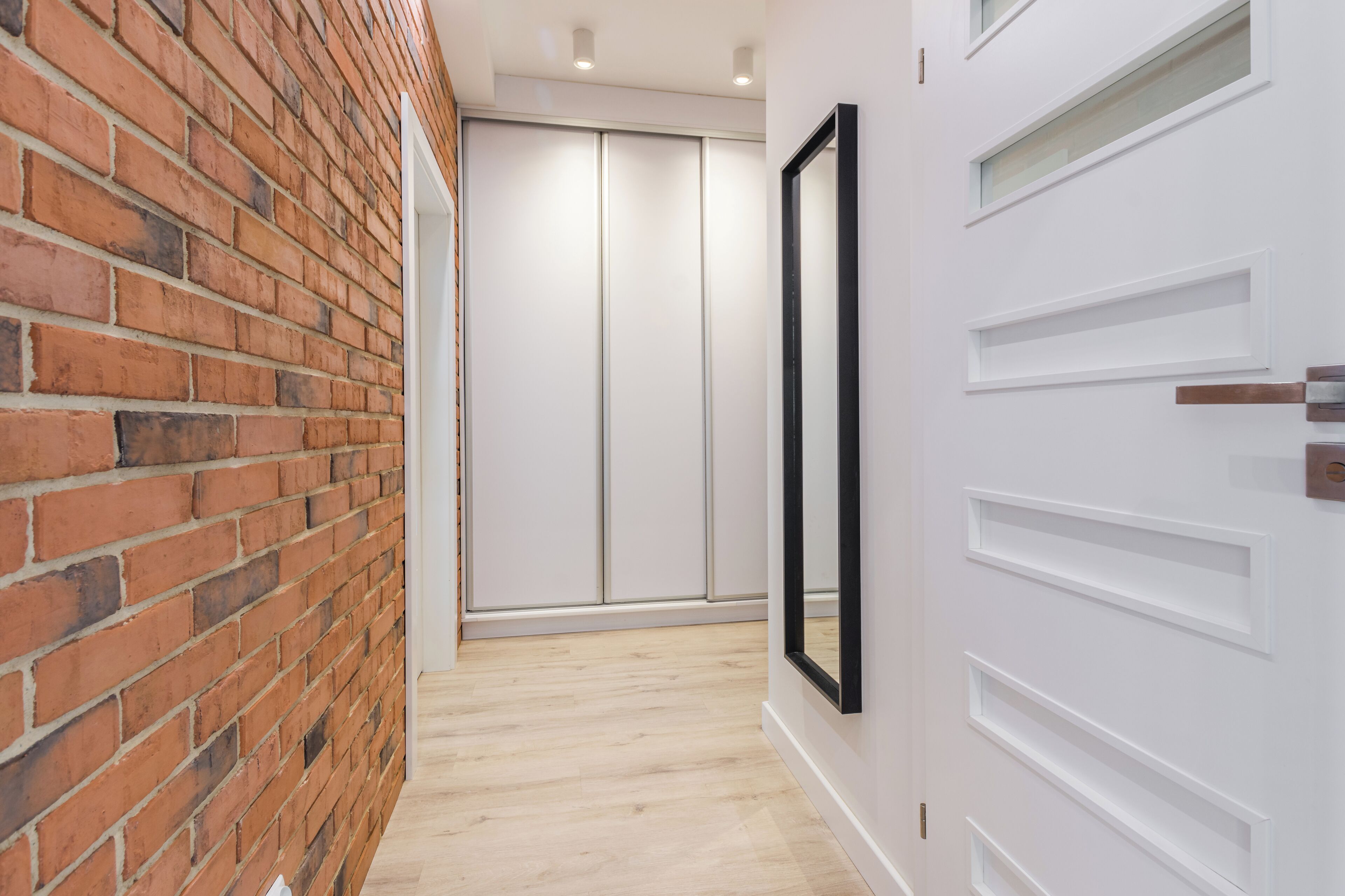 A hallway with a brick wall, a white door with a black frame, and a wooden floor.