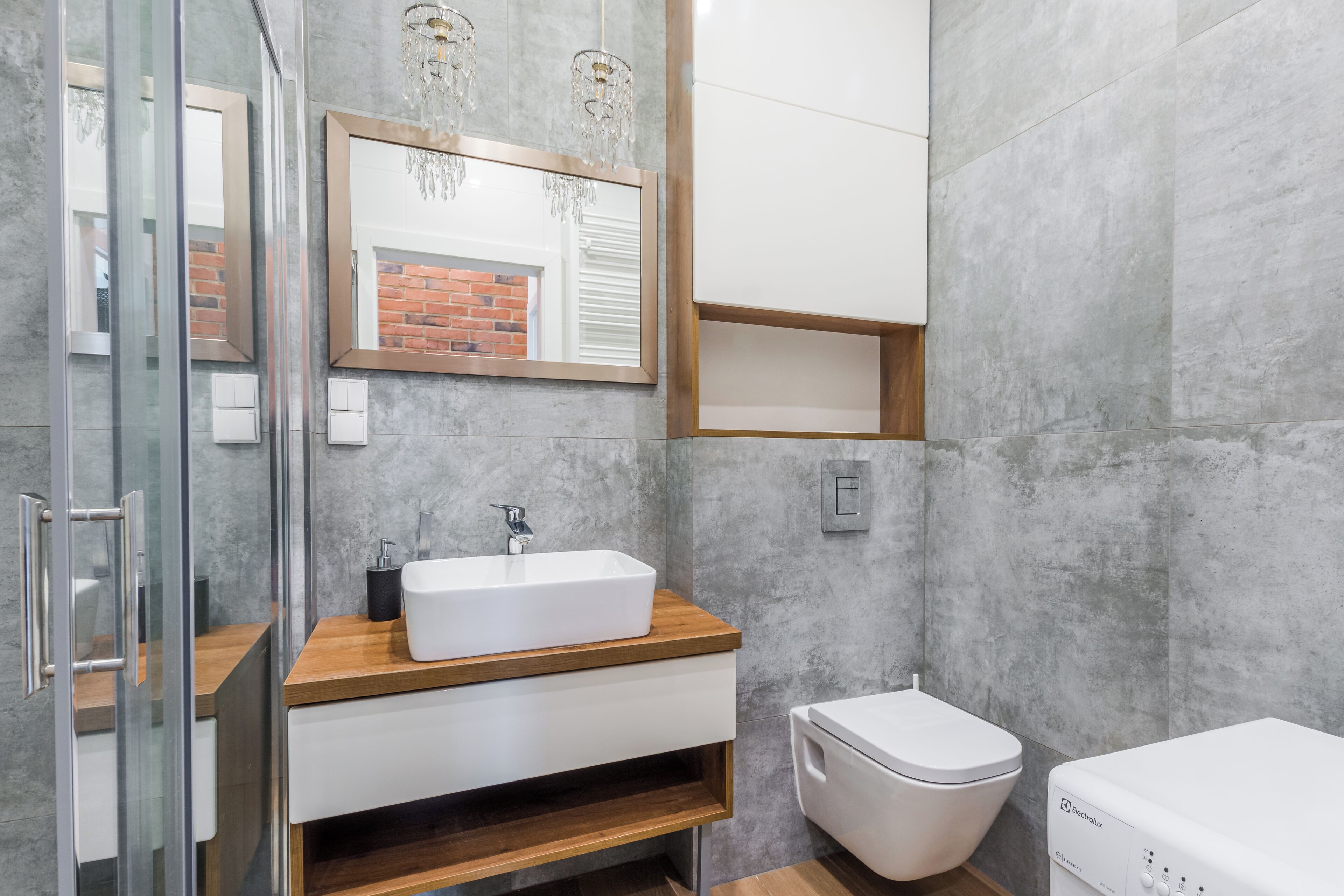 Un baño moderno con cabina de ducha de vidrio, tocador de madera con lavamanos blanco, inodoro blanco y espejo empotrado con candelabro.