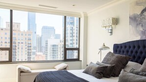 A bedroom with a large window, a bed with multiple pillows, a bench, and a wall-mounted light fixture. - Luxury Designer Penthouse 3803 | Pool | Gold Coast (Chicago)