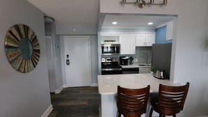 A modern kitchen with white cabinets, a stainless steel refrigerator, and a microwave. There is a dining area with two wooden chairs and a marble countertop.