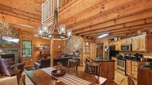 A cozy cabin interior with a wooden dining table, a fireplace, a kitchen with modern appliances, and a staircase leading to the upper level.