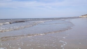 A beach with waves gently lapping onto the shore, a clear sky, and a distant shoreline with buildings. - Close to beach and amenities (Dives-sur-Mer)