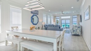 Un espacio habitable amplio con una gran mesa blanca, bancos, un sofá y un ventilador de techo. Un cartel en la pared dice "BEACH".