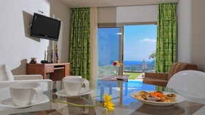 A hotel room with a dining table, chairs, a TV mounted on the wall, and a view of the ocean through large windows. - Villa Kazoulis (Kalathas)