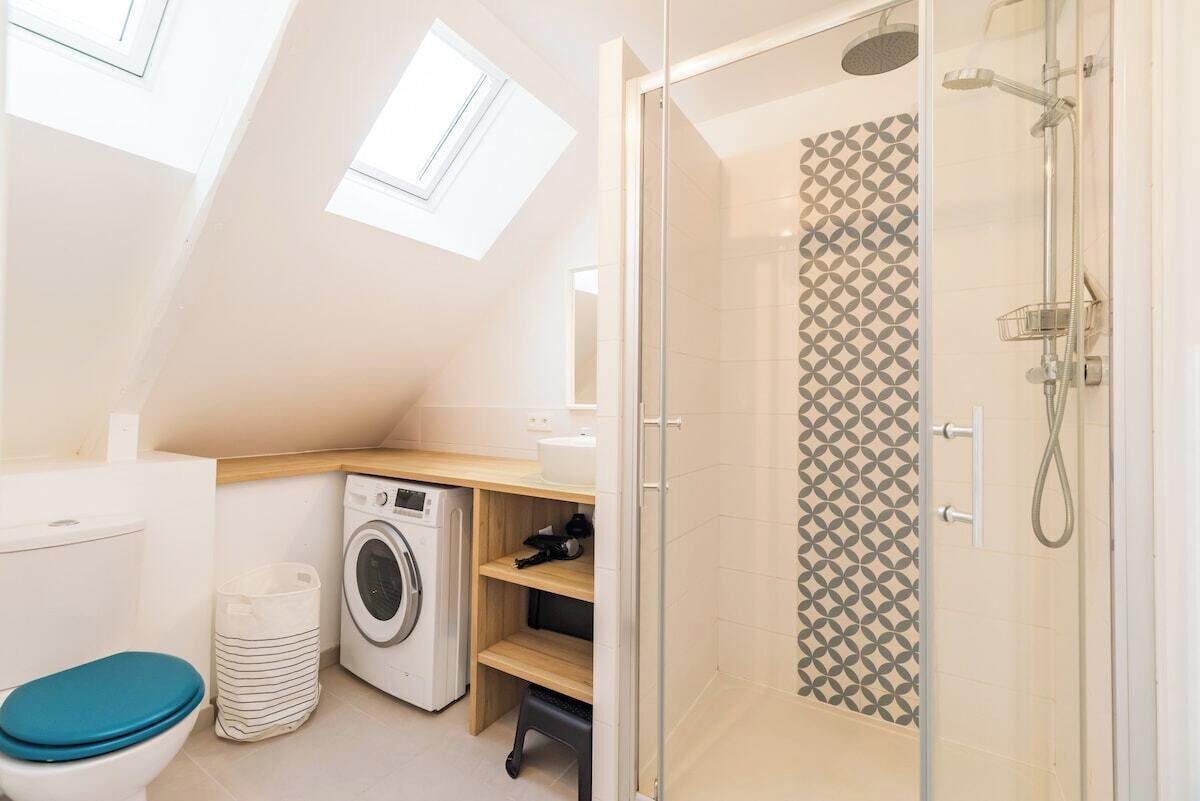 Un baño pequeño con tragaluz, ducha con azulejos estampados, lavadora e inodoro con asiento azul.