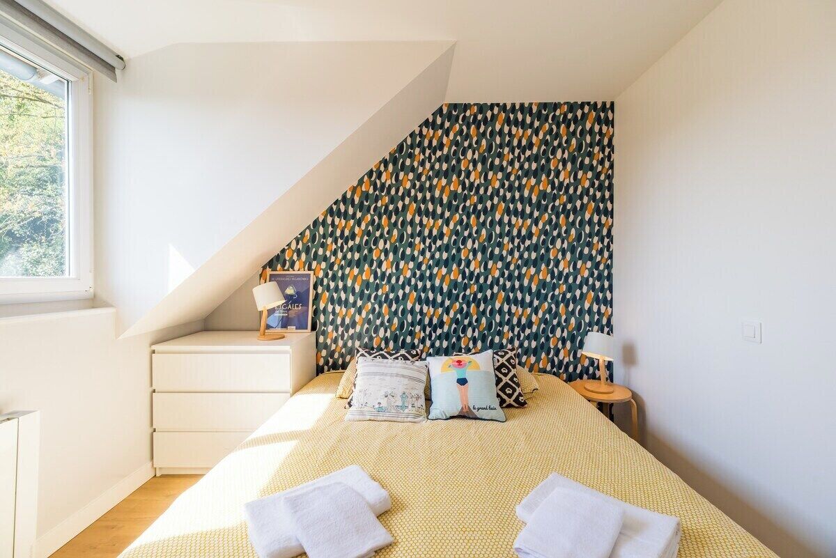 A bedroom with a sloped ceiling, a patterned wall, a bed with pillows, a bedside table, and a lamp.