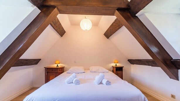 A bedroom with a sloped ceiling, wooden beams, a bed with white linens, two bedside tables with lamps, and a hanging light fixture. - Terrace, charm & nature in the heart of Lannion (Lannion)