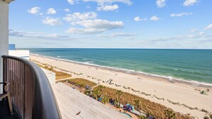 Une vue sur la plage depuis un balcon doté d’une terrasse en bois, avec une plage de sable et l’océan en toile de fond.