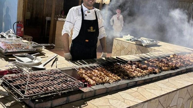 Un chef en una estación de parrilla con brochetas de carne, un extintor y una fuente de agua al fondo.