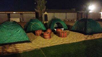 Four green tents arranged in a row on a grassy area with a wooden deck, surrounded by a wooden fence and a palm tree.