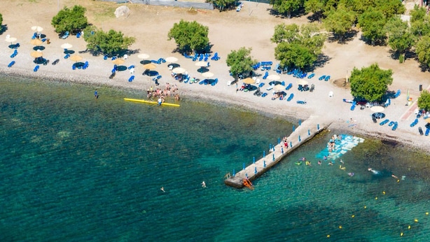 An aerial view of a beach with clear water, a pier, and numerous sun loungers and umbrellas.
