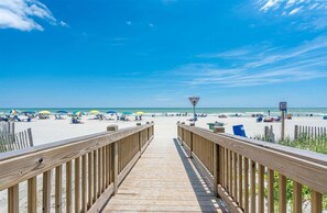 Ein hölzerner Steg führt zu einem Strand mit Sonnenschirmen und Menschen.