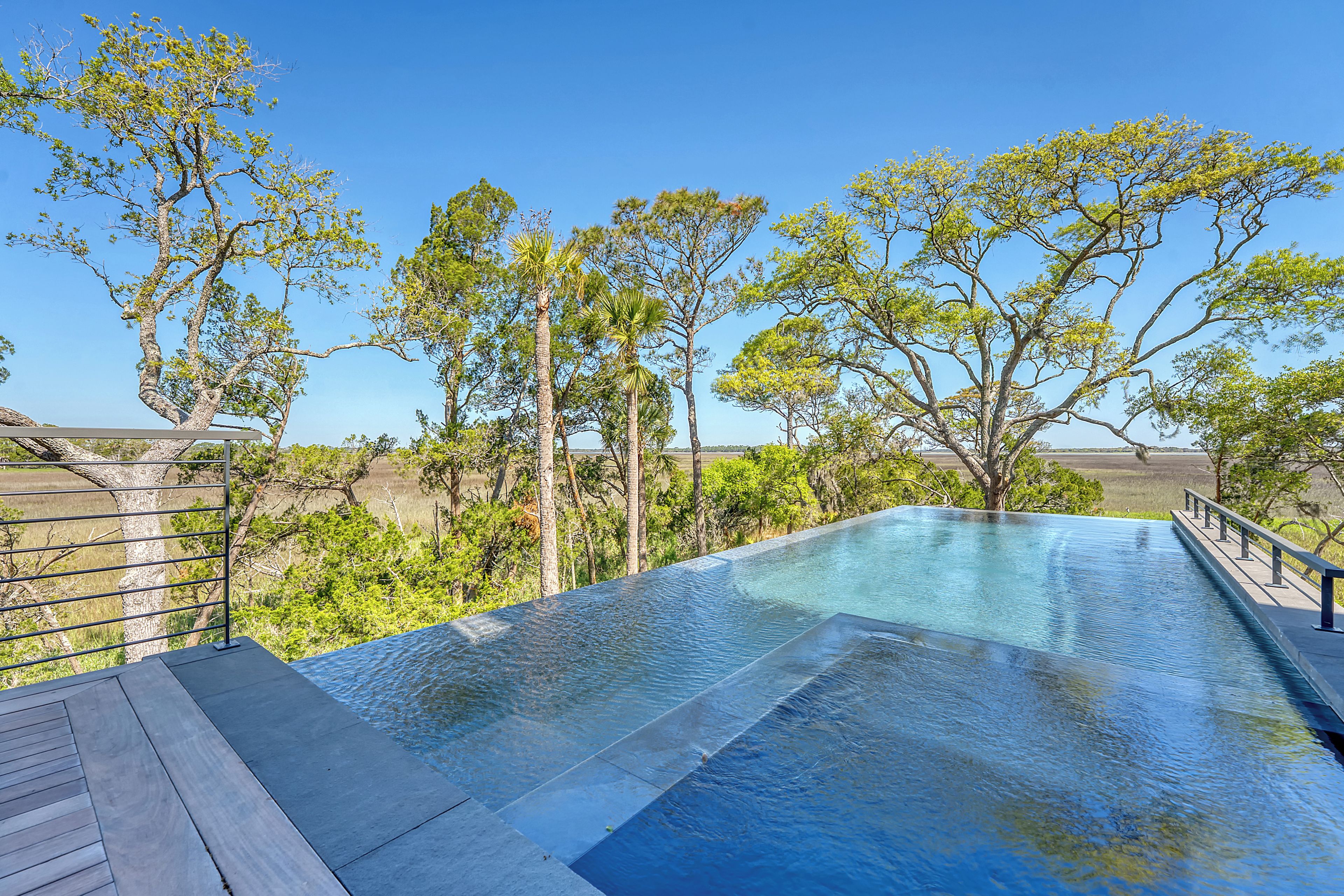 An infinity pool with a wooden deck and metal railing, surrounded by trees and a clear blue sky.