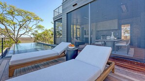 A modern outdoor deck with two lounge chairs, a small table with fruit, and a view of the pool and building interior.