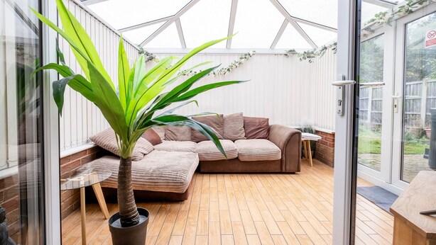 A conservatory with a cushioned seating area, a potted plant, and a wooden floor. - Colwell 4 Bedroom 3 Bathroom House (Derby)
