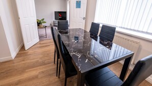 A modern dining area with a glass table and black chairs, a potted plant, and a TV in the background. - Colwell 4 Bedroom 3 Bathroom House (Derby)