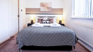A neatly made bed with a patterned bedspread, two bedside lamps, and a wall-mounted artwork above the bed. - Colwell 4 Bedroom 3 Bathroom House (Derby)