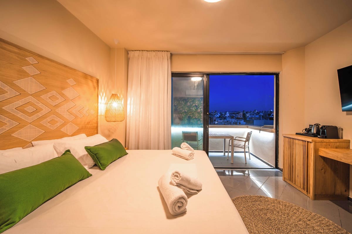 Habitación de hotel con una cama grande, almohadas verdes, cabecera de madera, escritorio con cafetera y vista nocturna de la ciudad.