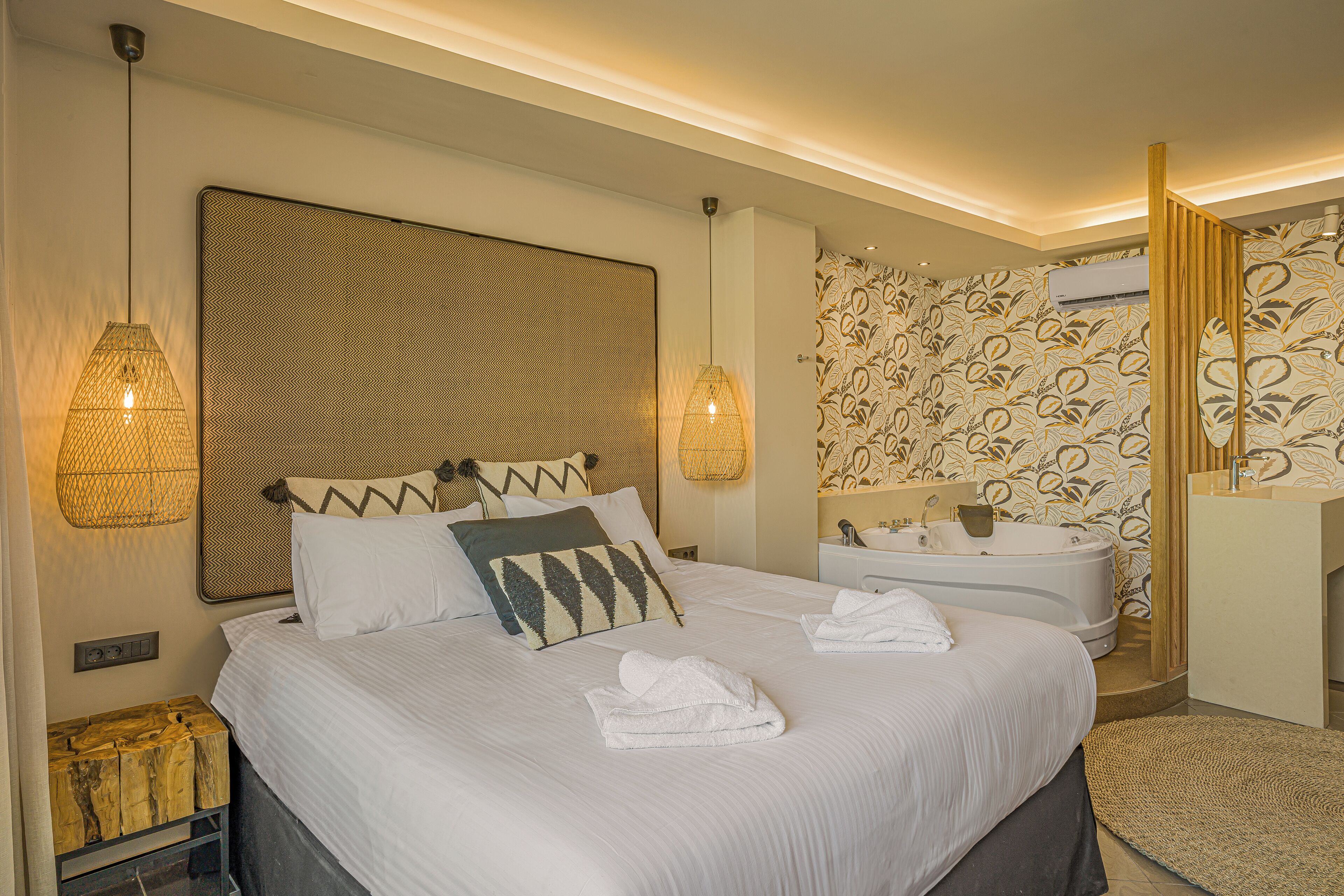 Une chambre d’hôtel avec un grand lit, deux lampes fixées au mur, une tête de lit, un climatiseur mural, une salle de bain avec baignoire et un miroir.