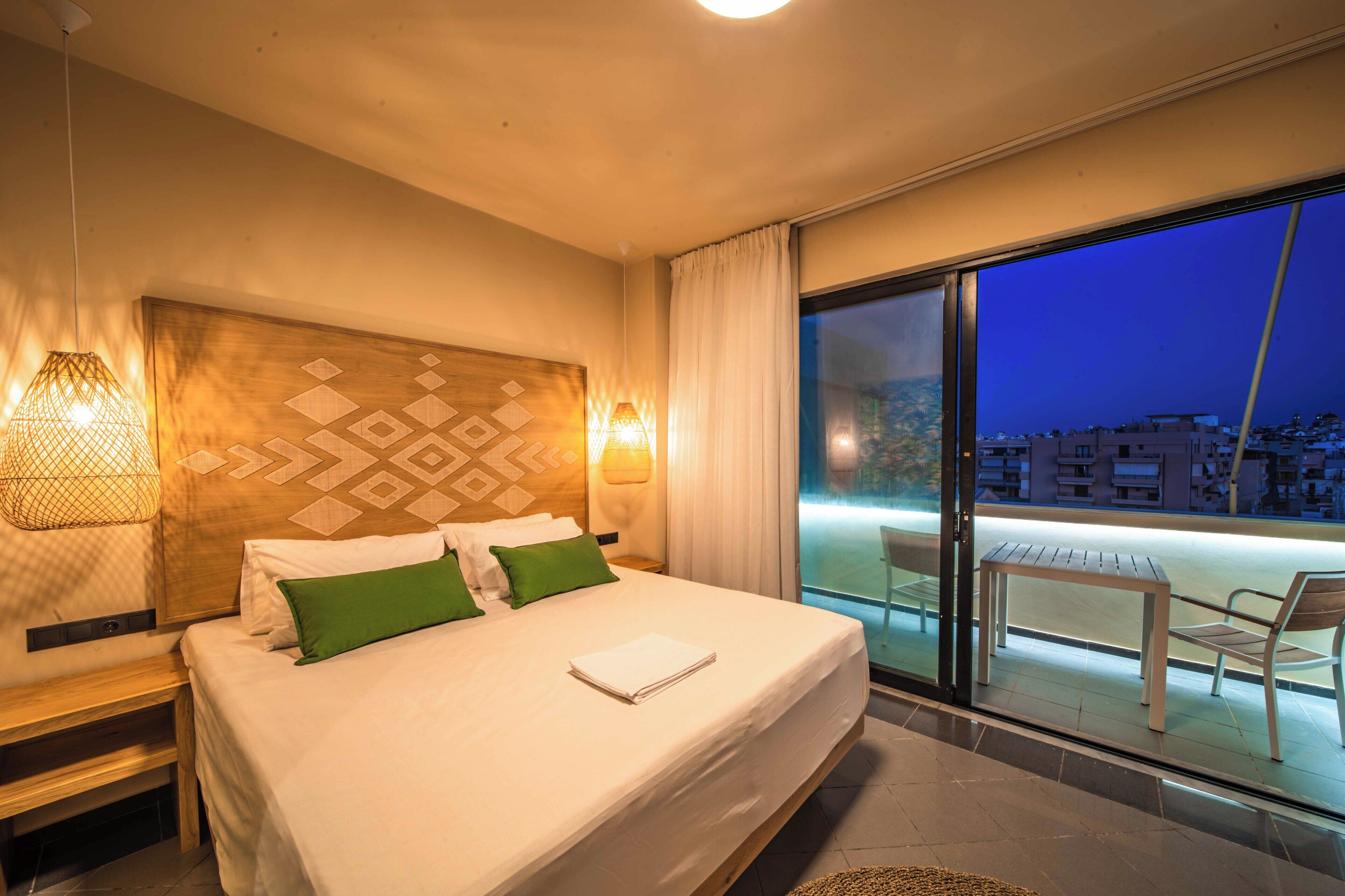 A hotel room with a large bed, two bedside lamps, a wooden headboard, a balcony with a table and chairs, and a view of the city at night.