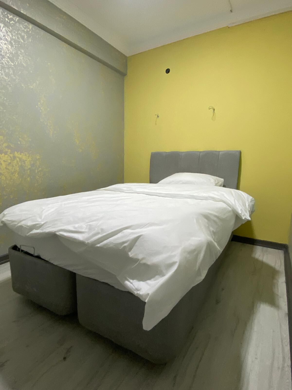 Una cama individual con cabecera gris en una habitación con paredes amarillas y suelo gris.