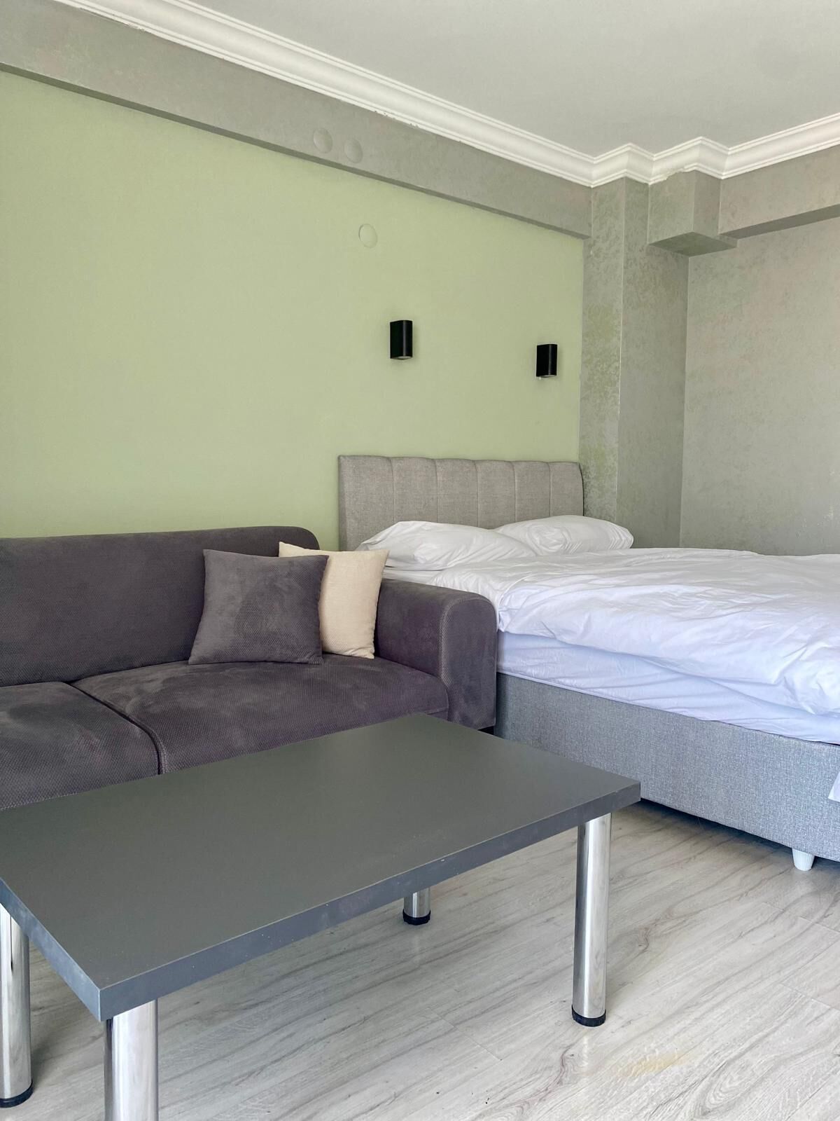 A modern hotel room with a grey sofa, a small table, a bed with white linens, and two wall-mounted light fixtures.