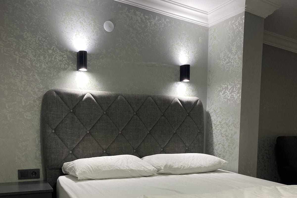 Un dormitorio con cabecera capitonada, dos lámparas de noche, una cama con ropa blanca y una mesita de noche con cajones.