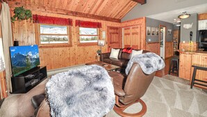 Interior acogedor de una cabaña con paredes de madera, un sofá de cuero, un sillón cubierto de piel y un televisor mostrando un paisaje montañoso.