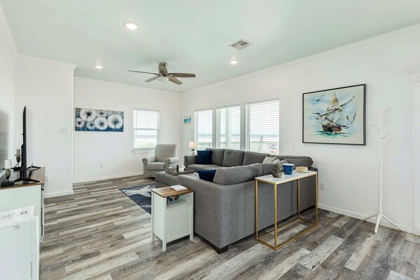 A modern living room with a gray sectional sofa, a white side table, a small white cabinet, and a blue rug. There are two windows with blinds, a ceiling fan, and a framed painting of a sailboat. - Spacious Beachfront Home in Surfside Texas (Surfside Beach)