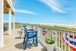 A wooden deck with blue chairs and tables, overlooking a beach with flags. - Spacious Beachfront Home in Surfside Texas (Surfside Beach)