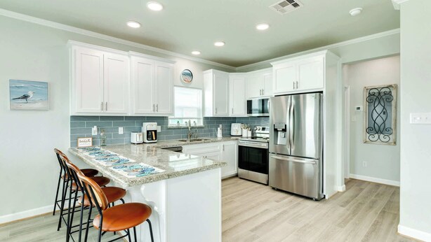 A modern kitchen with white cabinets, a stainless steel refrigerator, a microwave, and a coffee maker. There are three bar stools at the island. - Spacious 3 Bedroom Surfside Home Near The Jetties (Surfside Beach)